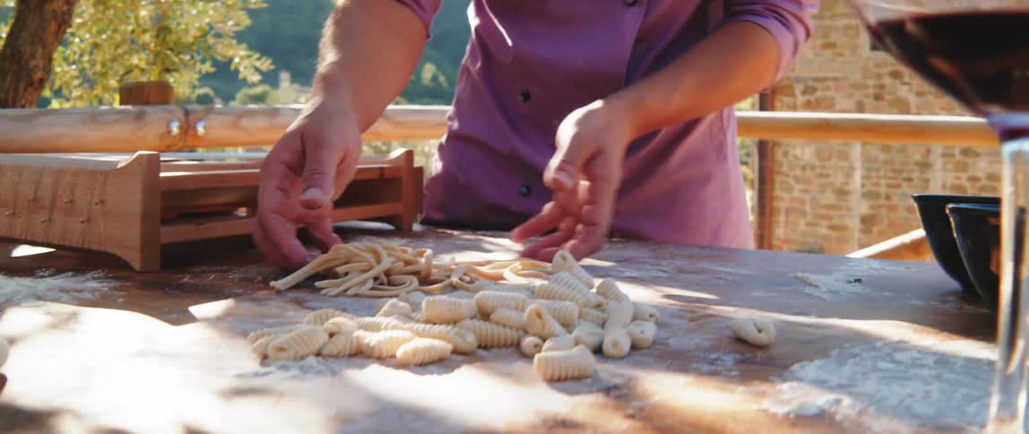 A person in a purple chefs jacket preparing pasta on a wooden table outside