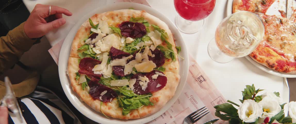 Person sitting at a table with pizza topped with cheese and green leaves served on a plate and drinking a pink drink