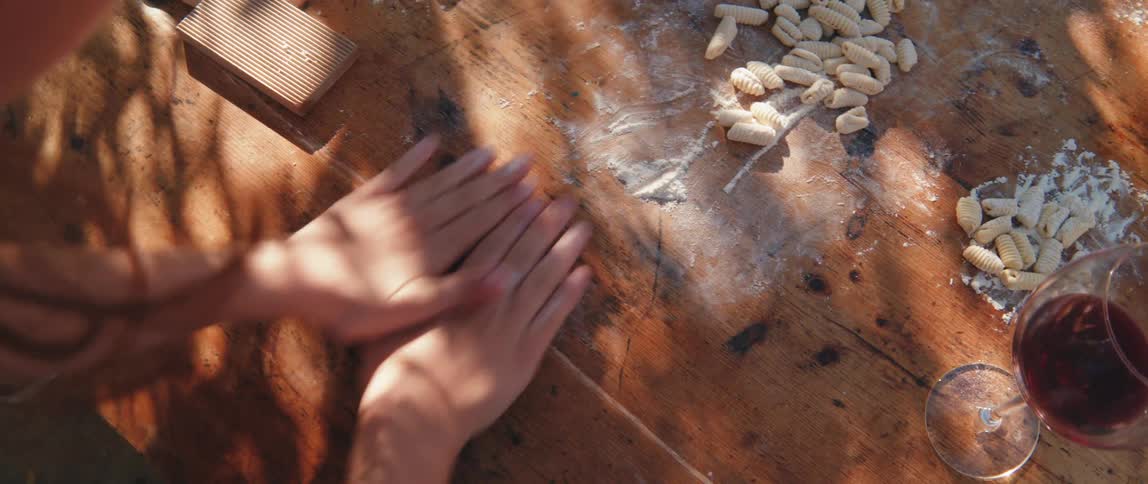 Hands of a person making pasta dough rolls on a wooden table with a glass of wine next to it
