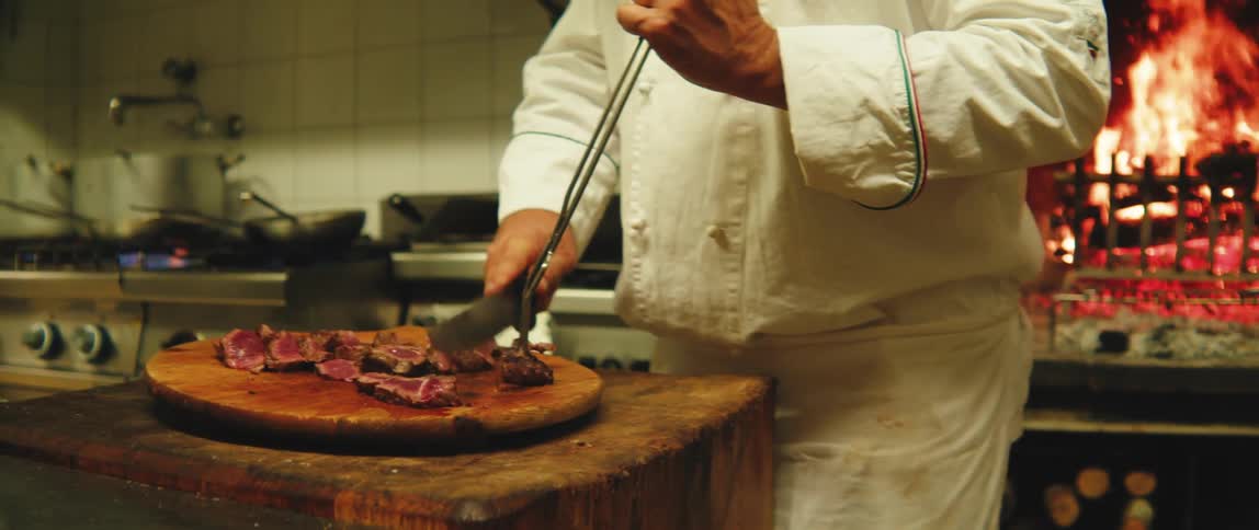 Man in an apron inside a restaurant kitchen cutting roast meat on a cutting board