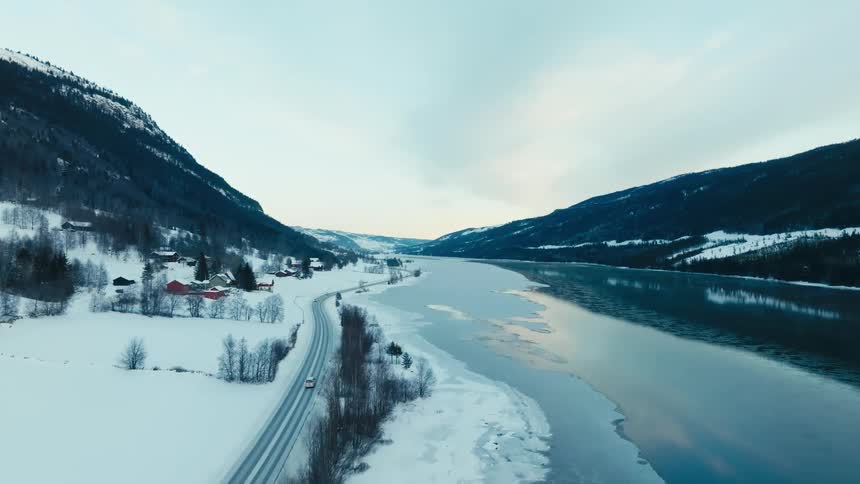 Automobiles travel on a snow covered road near houses along an ice covered river in a forested mountain valley under a cloudy sky