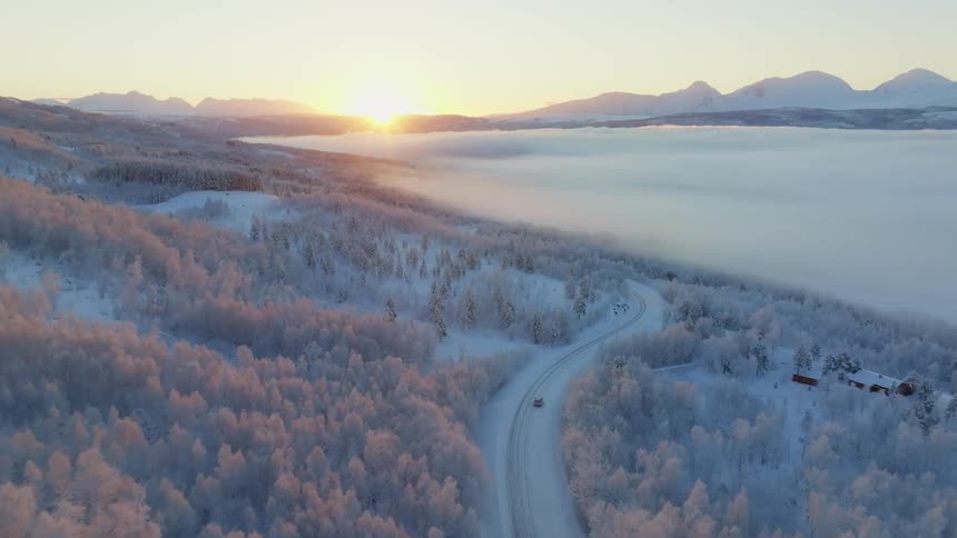 Aerial view of a natural icecovered environment in the mountains trees and a lake with a vehicle passing on the road and the sunrise on the horizon