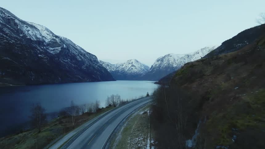 Aerial of cars driving on highway near the lake with high snowy mountains in the background