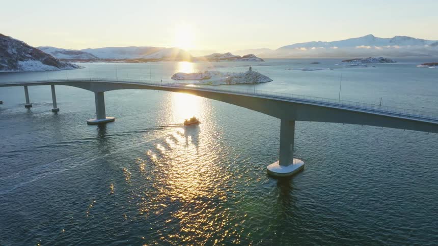 Boat crossing under bridge leaves wake through water reflecting sunlight on winter day