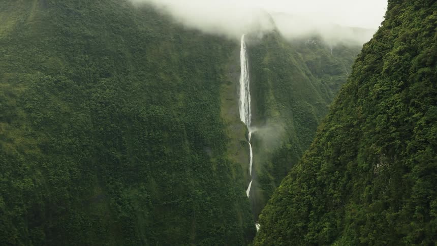 Waterfall flows down steep mountainside into narrow valley between green mountains under low lying clouds
