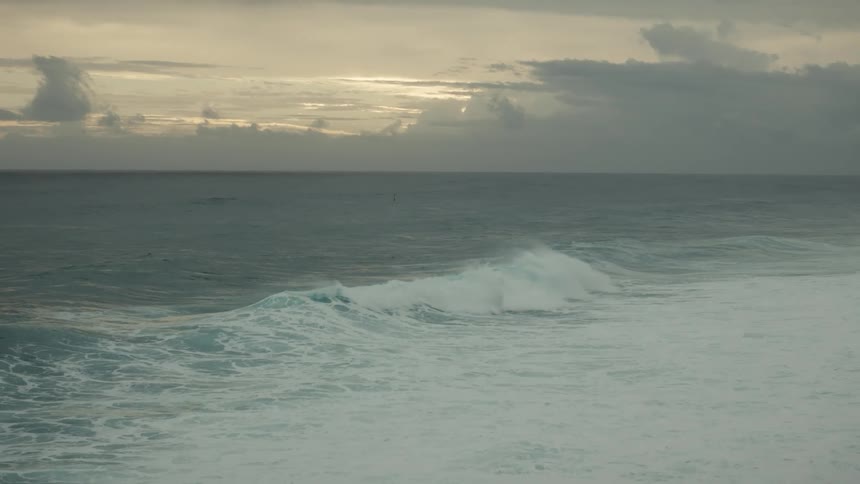 A seascape with waves rolling in under a cloudy sky at dusk