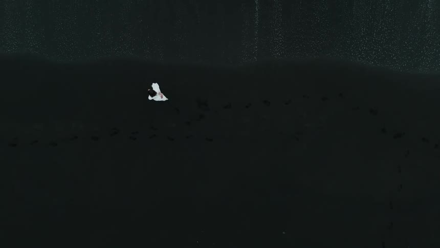 A woman in a white colored dress twirls and walks on a dark sandy beach
