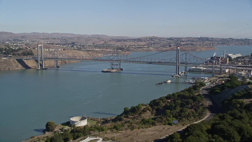Transmission towers stand on hill overlooking river near businesses across from residential neighborhoods