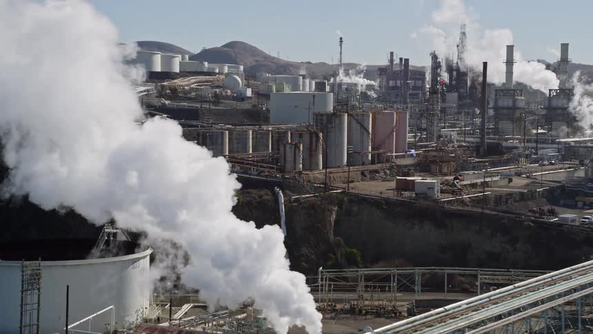 An aerial view of the sea and in front of a large industry with factory buildings releasing white smoke