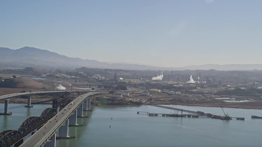Aerial panorama of an extensive bridge under the sea with vehicle traffic that gives access to a city with factories and urban areas and the silhouette of mountains on the horizon