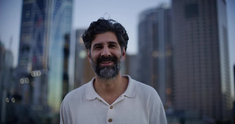 A bearded man in a white shirt is smiling and behind him are several tall buildings