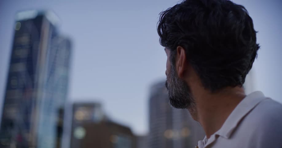 A bearded man is looking ahead where there are several tall buildings in a city on a day with a blue sky