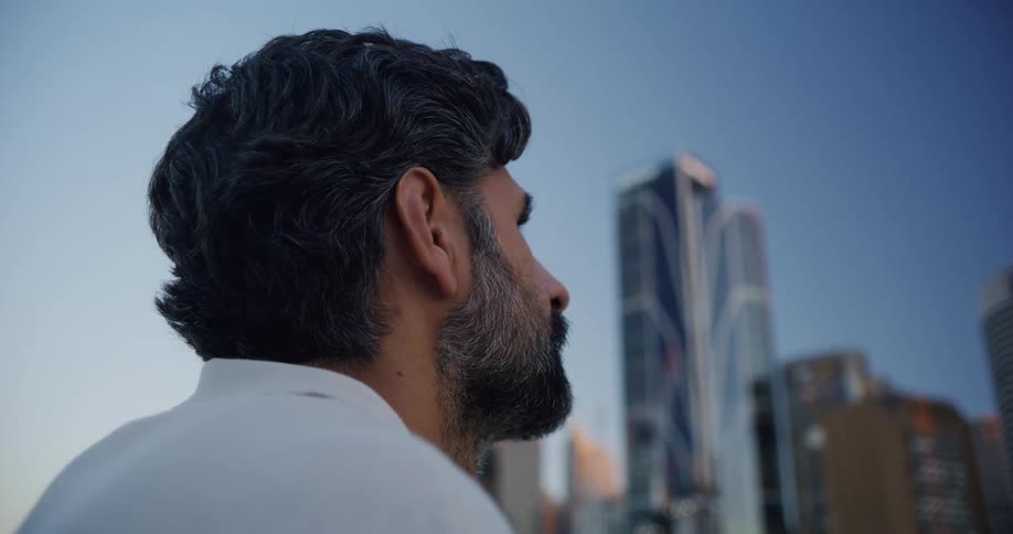 A bearded man is looking ahead where there are several tall buildings