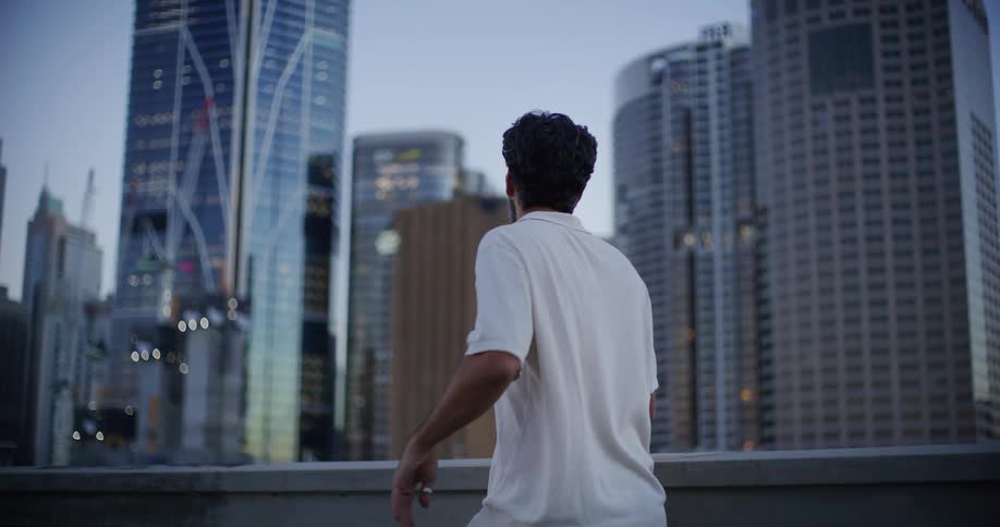 A bearded man in a white shirt is looking to the side and then turns his back and looks at an area with several tall buildings in a city