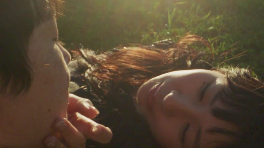 Couple in love looking at each other while the woman passes her hand on her partners face and they are lying on a grass with a reflection of sunlight