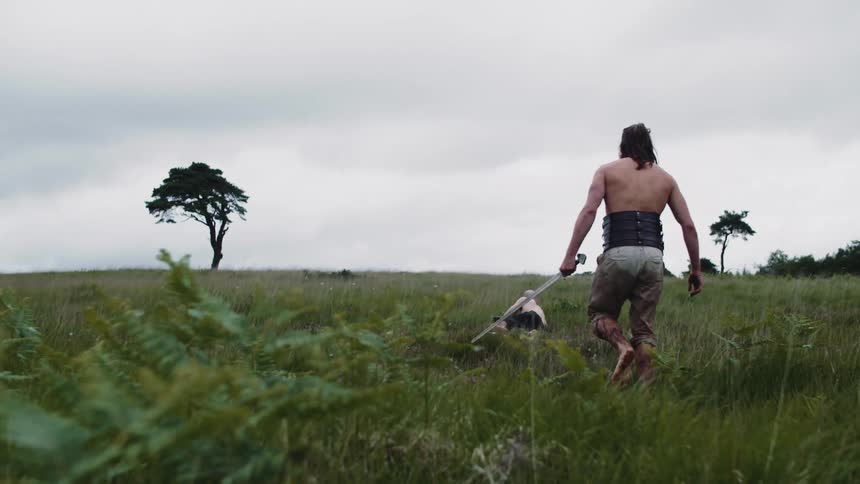 Man carrying a sword walks after man crawling across a field on an overcast day