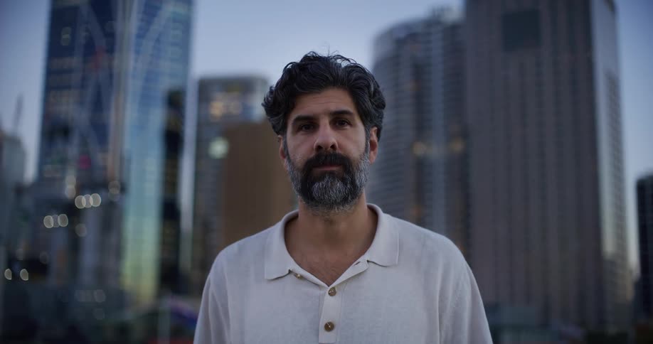 A bearded man in a casual collared shirt stands before a city skyline