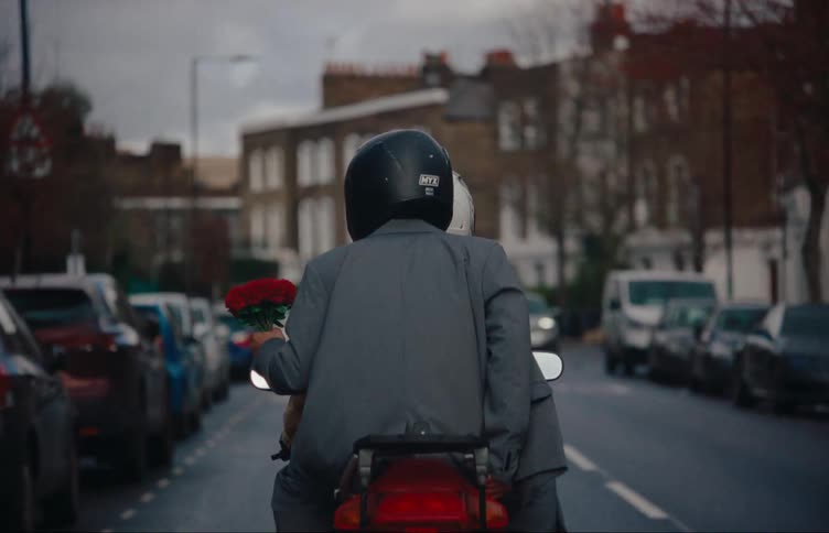 A person in a dark helmet and jacket rides a scooter down a street with parked cars