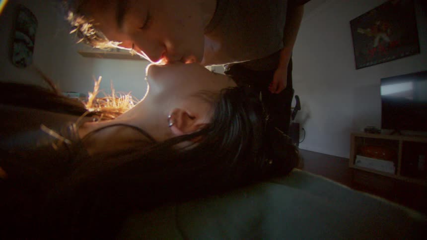 A young woman lays on a bed her boyfriend leans down giving her a kiss