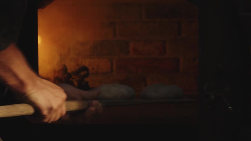 Male baker places large loaves of bread into smoking wood oven