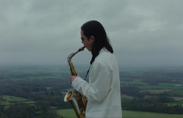 A man in white clothes and sunglasses is playing the saxophone and behind him there is a huge field with trees