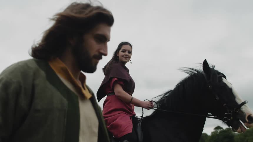 Man walking next to a woman who is walking a black horse in an open field on a cloudy day