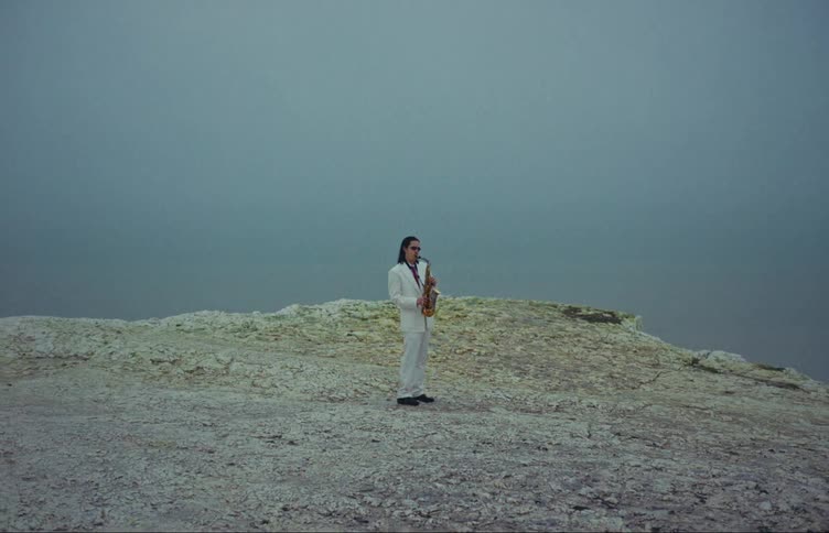 Man in a gray suit playing the saxophone on a rock outdoors on a cloudy day