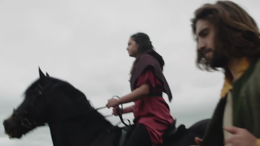 Portrait of a medieval man walking next to a woman who is riding a horse in an outdoor setting on a cloudy day