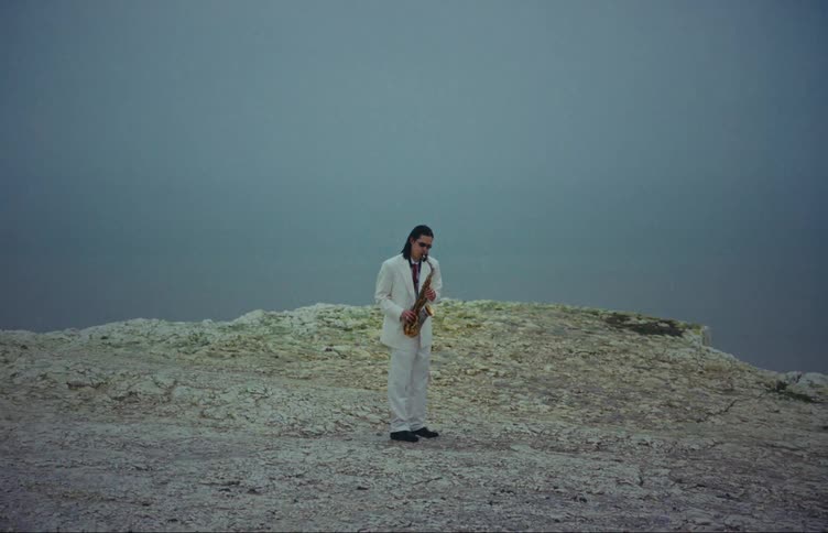 Man in white suit playing saxophone on a cliff