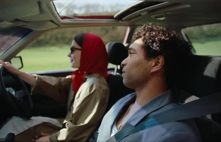A woman with sunglasses and a red hood is driving a car and next to her there is a man and the sunroof of the car is open