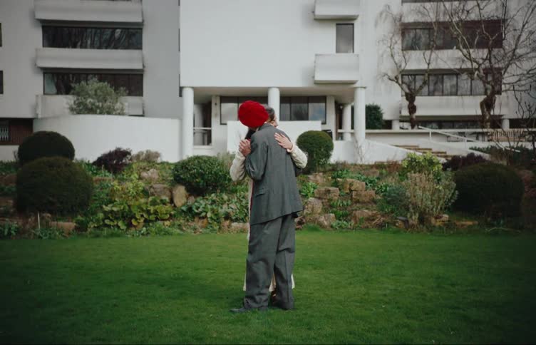 An elderly couple dances together on the lawn in front of an apartment building
