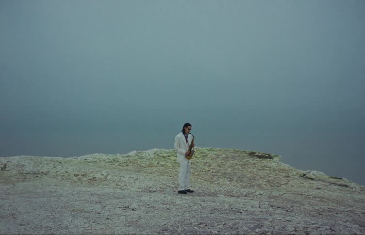 A man in a white suit plays a sax on a mountaintop