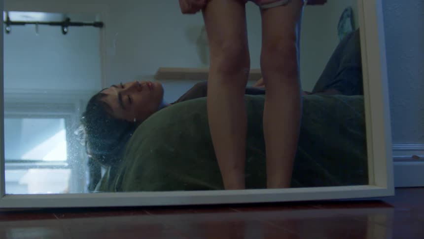 Man lying in bed and next to him his girlfriend taking off her pink shorts in front of the mirror