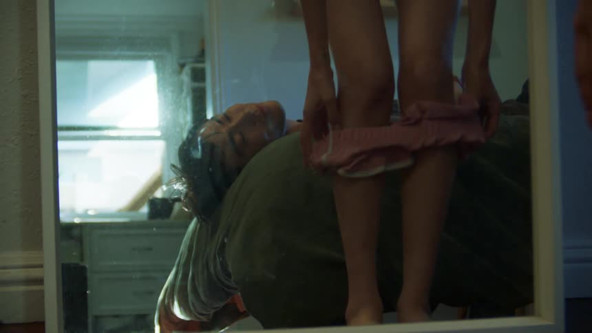 Man lying in bed and his girlfriend changing in front of the mirror taking off part of her clothes