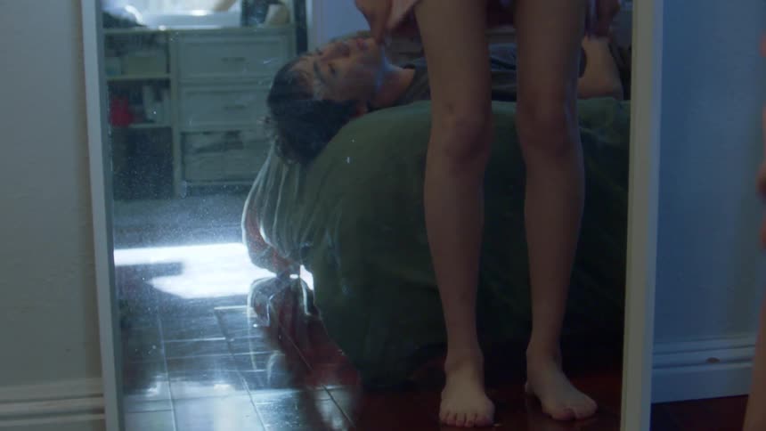 Man lying in bed and his girlfriend taking off her shorts in front of the mirror