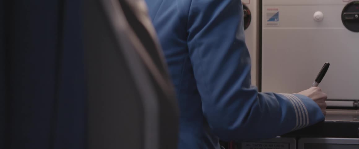 A woman in a blue outfit has her back turned and is sitting and writing something using a black pen