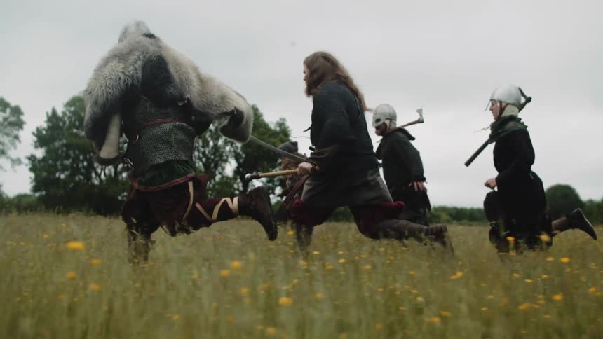 A group of people dressed as ancient medieval soldiers  run across a field with weapons drawn