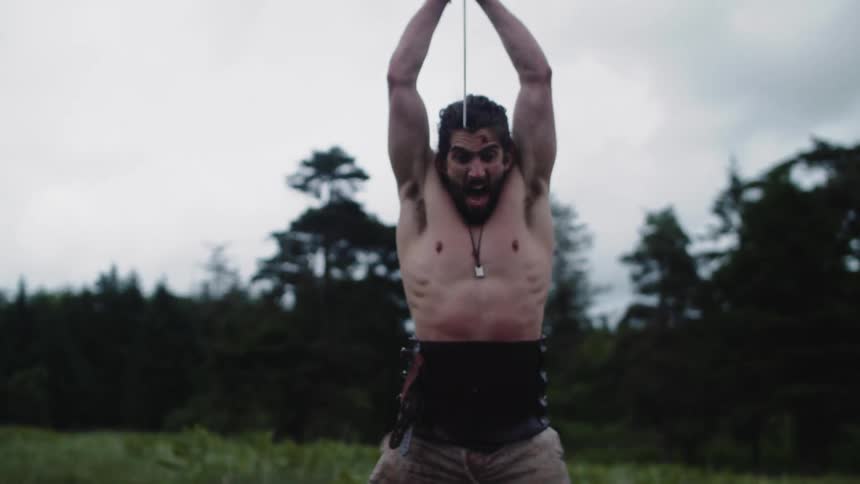 Viking man with a sword killing a man who is lying on a lawn in a field on a cloudy day