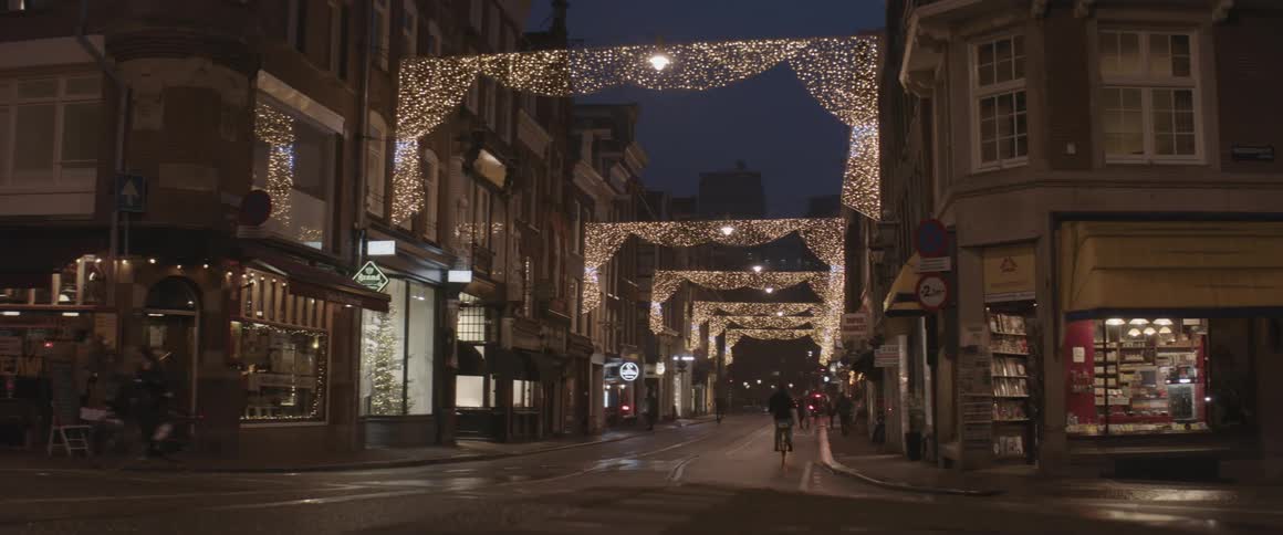 An asphalt street with decorative lights connecting buildings on both sides and cars and motorcycles passing through the street