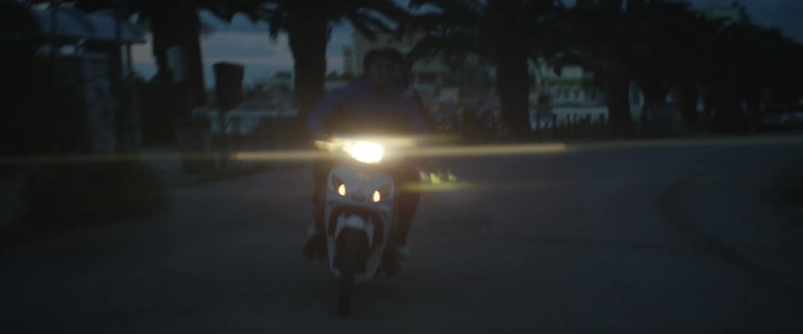 A man and a passenger ride a motorcycle through an urban, coastal community at dawn