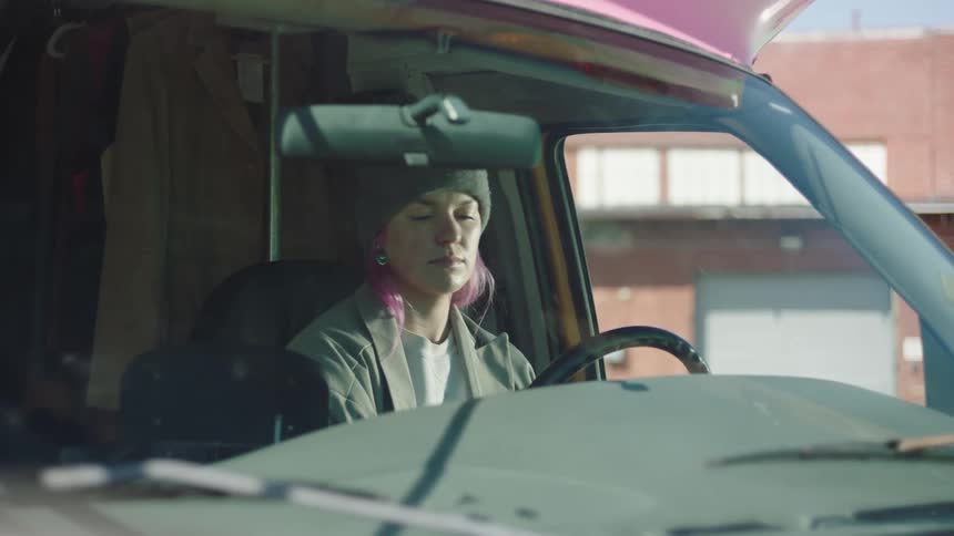 A female wearing a beanie sits in the driver seat of a shuttle bus and opens its door