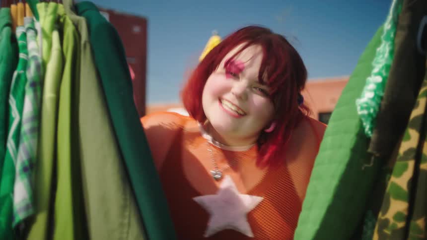 A woman in fantasy makeup going through a clothes rack then spreading the clothes and peeking through laughing