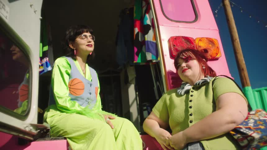 Two models with one of them sitting on the back of a pink bus while wearing trendy green colored clothes