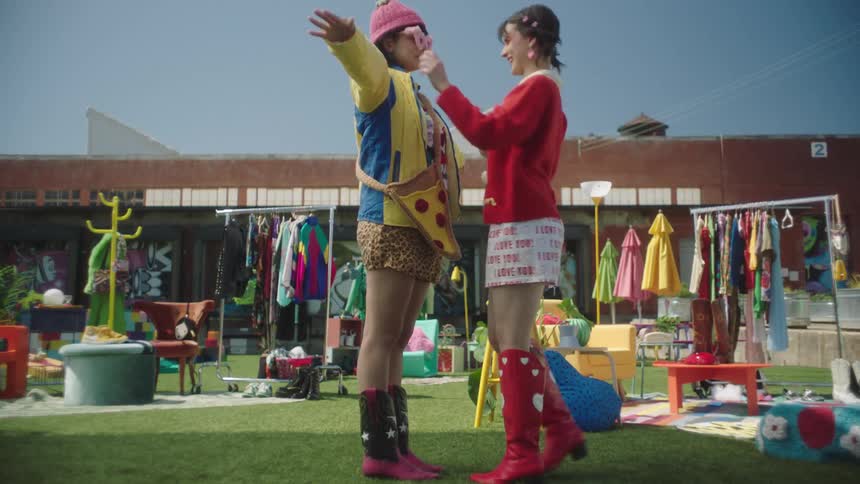 Woman putting colored sunglasses on a model who is standing on the grass and wearing a colorful outfit