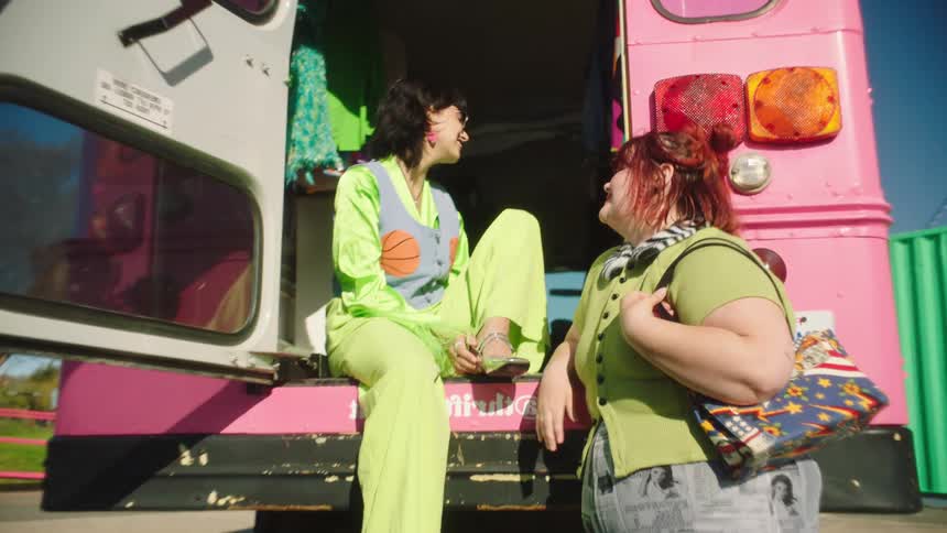 Two female models talking and smiling while posing in light green clothes and on a pink transport outside