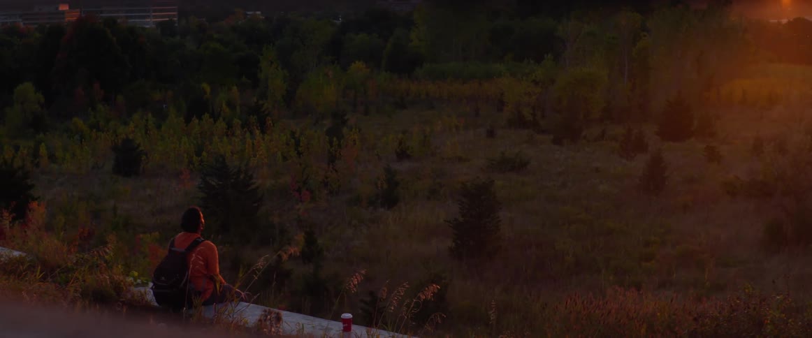 A backpacker sitting on a fallen tree trunk looking at a forest near a city