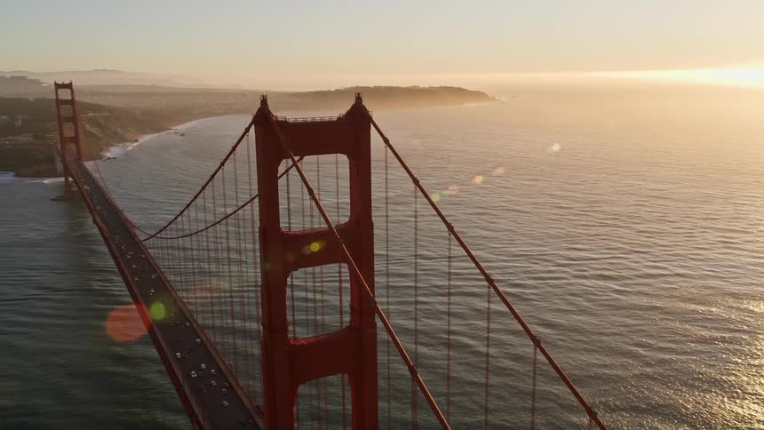 Aerial panorama of the golden bridge with vehicle traffic on the edge of the mountains and the sun on the horizon