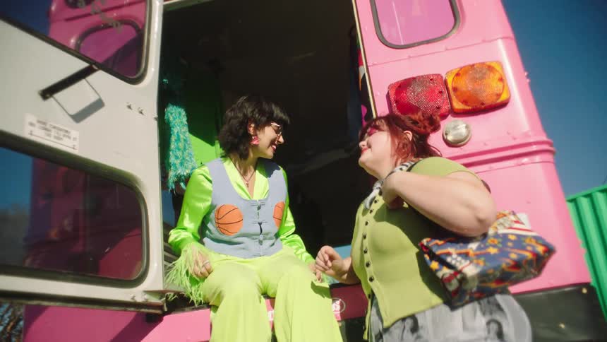 Two smiling women with one of them sitting in the back of a truck wearing fashionable greenish clothes