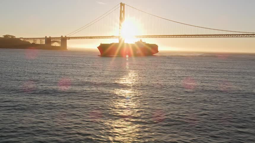 Aerial panorama of a cargo ship traveling near the golden bridge with sun reflection on the horizon
