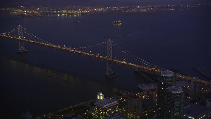 An aerial view of a huge bridge with many vehicles passing by at dusk and the bridge is suspended over a bay